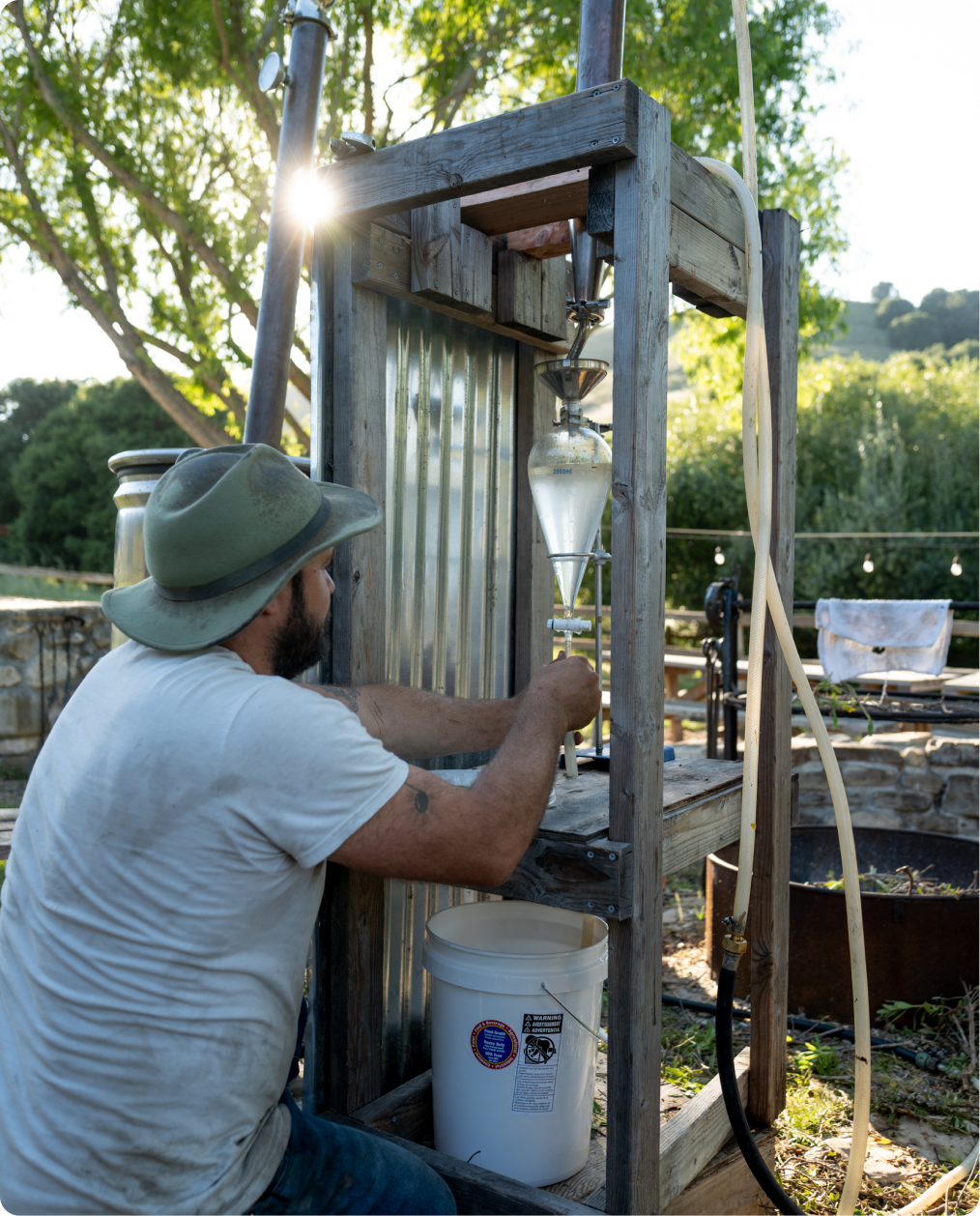 A man in a hat operates a makeshift distillation apparatus.